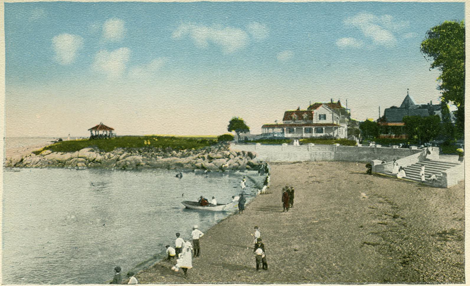 People walk and stand along a sandy beach bordered by rocks and water, with several large houses and a gazebo visible in the background under a blue sky with scattered clouds.