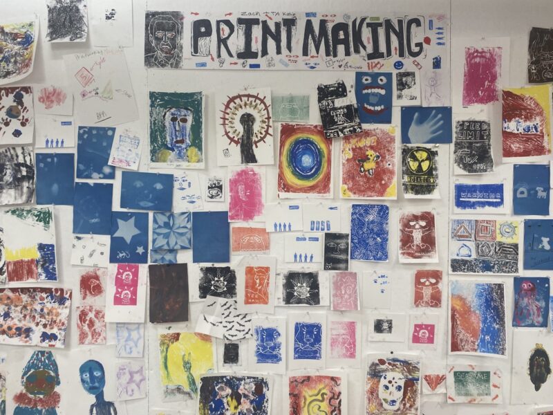 A wall is covered with various colorful printmaking artworks and sketches, with a large sign at the top reading "PRINT MAKING" in bold black letters. The prints show abstract designs, faces, and different shapes.