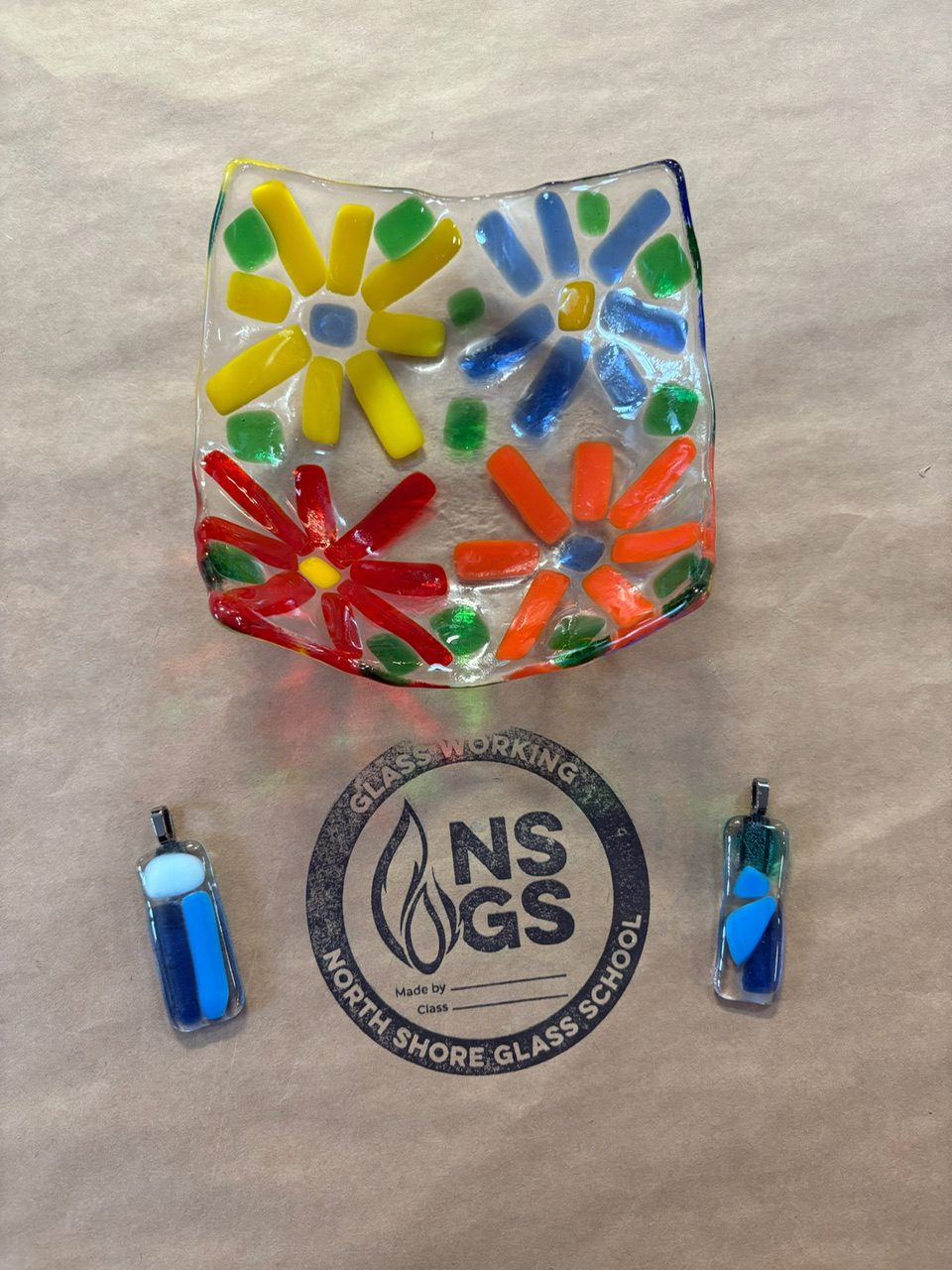 A square fused glass plate with colorful flower designs and two small glass pendants sit on brown paper with the North Shore Glass School logo printed on it.