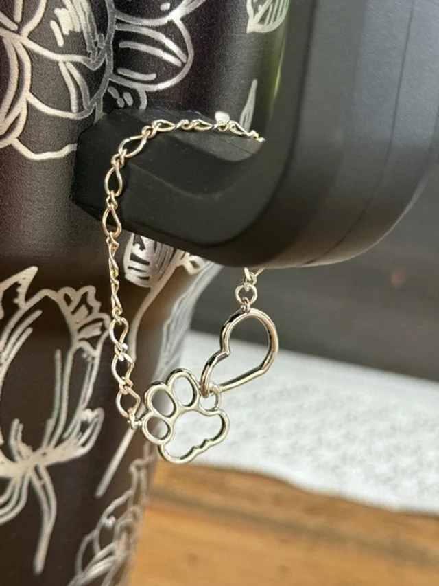 A silver chain with a paw print and heart-shaped charm, inspired by mixed metals permanent jewelry, is hanging from the handle of a black and white floral-patterned mug.
