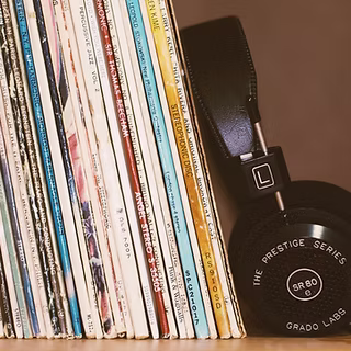 A stack of vinyl records stands upright next to a pair of black over-ear headphones labeled "The Prestige Series SR80 Grado Labs," evoking the spirit of band practice against a neutral background.
