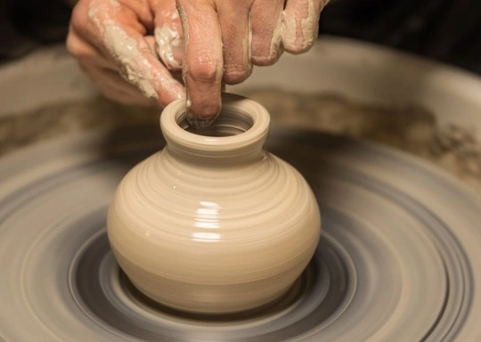 A person’s hands shaping a small clay pot on a spinning pottery wheel, expertly demonstrating wheel throwing with wet clay visible on their fingers as the pot takes form.