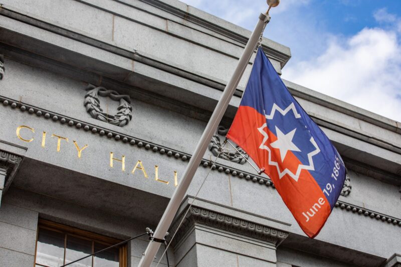 A Juneteenth flag with red, white, and blue colors and the date "June 19, 1865" flies at half-staff in front of a gray stone building with "City Hall" inscribed in gold letters.