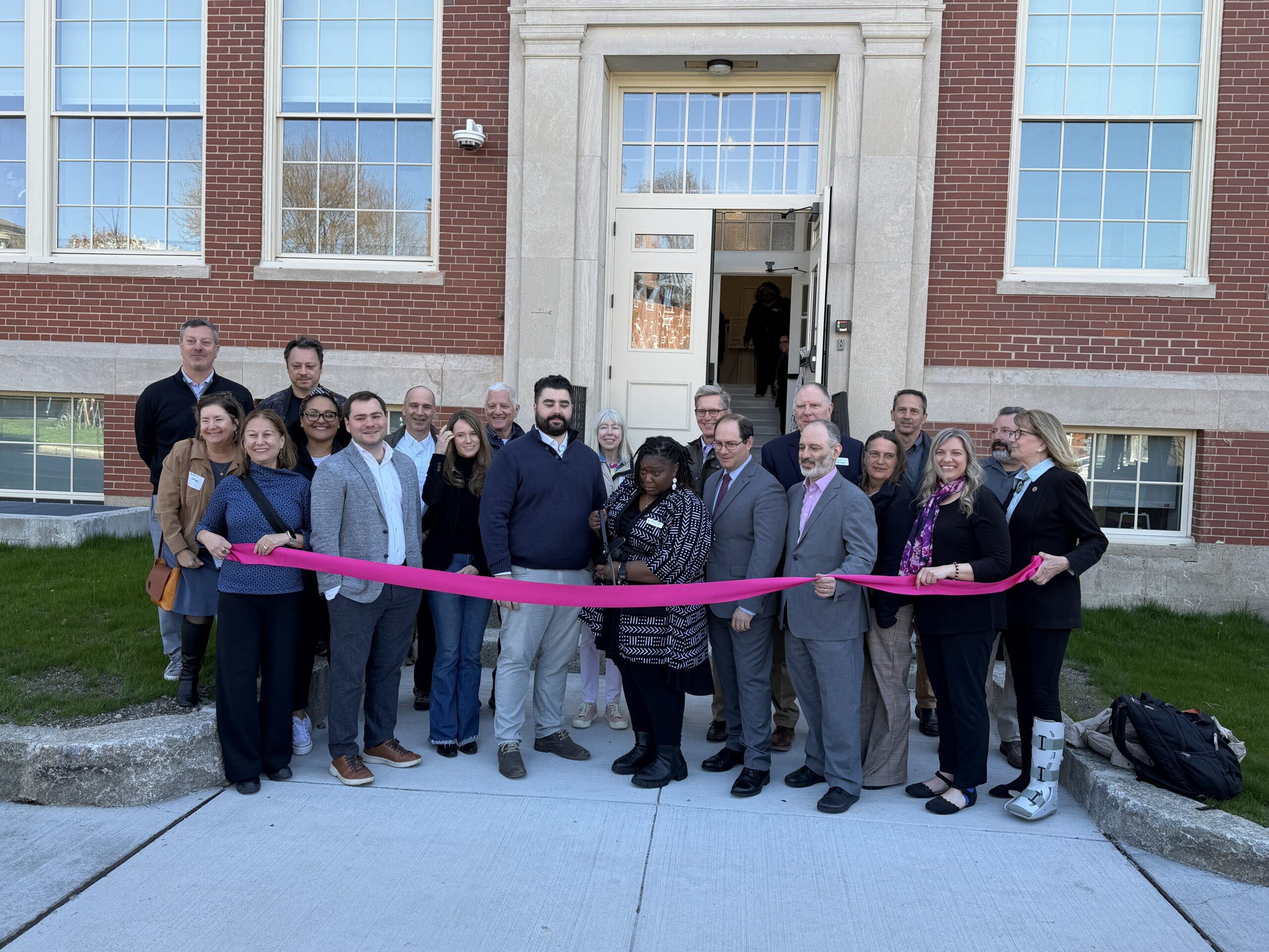 A group of about 20 people stands outside Hawthorne Lofts in Salem, holding a long pink ribbon during a ribbon-cutting ceremony for new affordable artist housing. Most are dressed in business or business-casual attire.