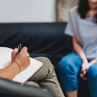 A person sits on a couch holding a notebook and pen, facing another person across from them. The image suggests a conversation, possibly a drop-in counseling session for survivors of sexual assault.