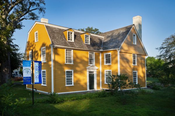 A two-story historic yellow house with white trim and a gambrel roof stands on a grassy lot. A blue sign reading "Historic" is posted near the building. The sky is clear and trees surround the area.
