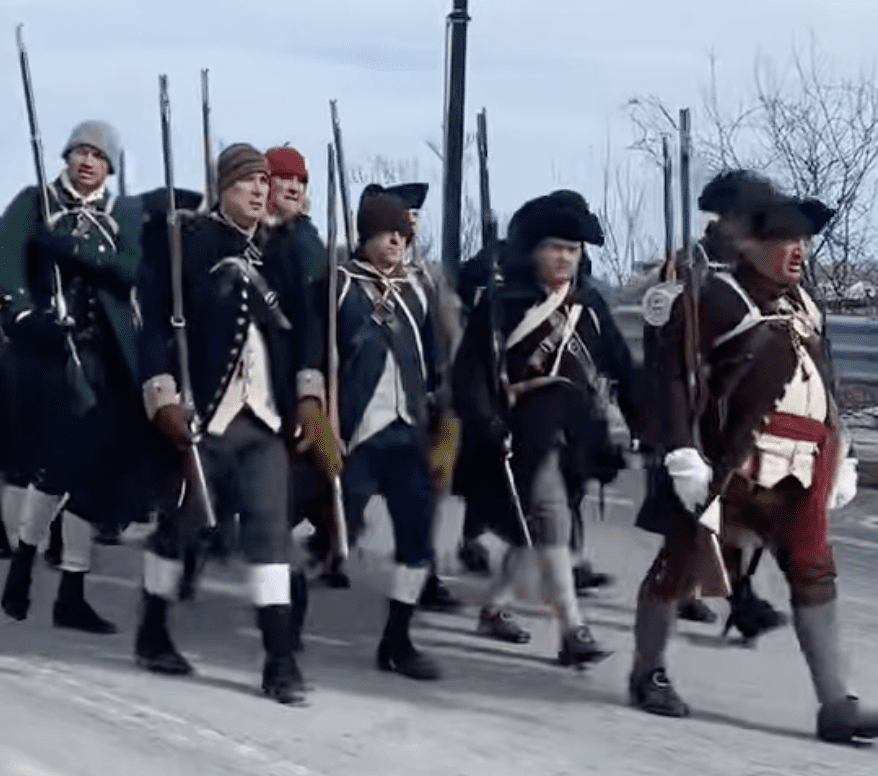 A group of men in historical military uniforms march in formation on a paved road, carrying muskets over their shoulders. The background shows leafless trees and a cloudy sky.