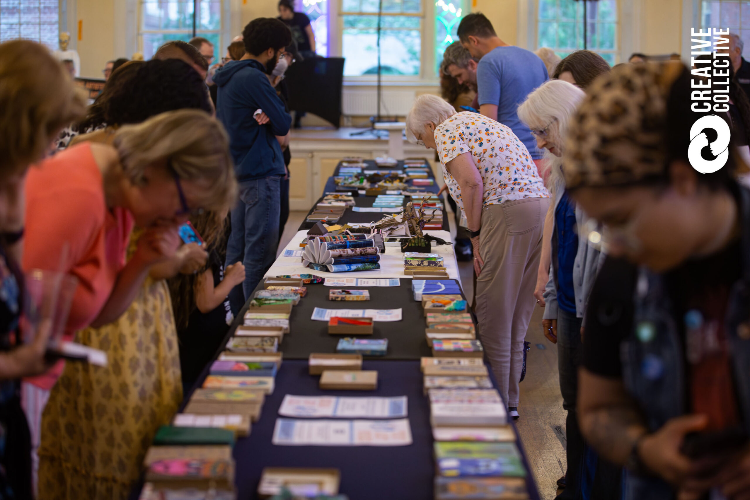 People stand around a long table covered with books and pamphlets at the Salem Arts Festival Tiny Art Spree, closely examining the items. The setting appears to be an indoor event, with a "Creative Collective" logo on the right side of the image.