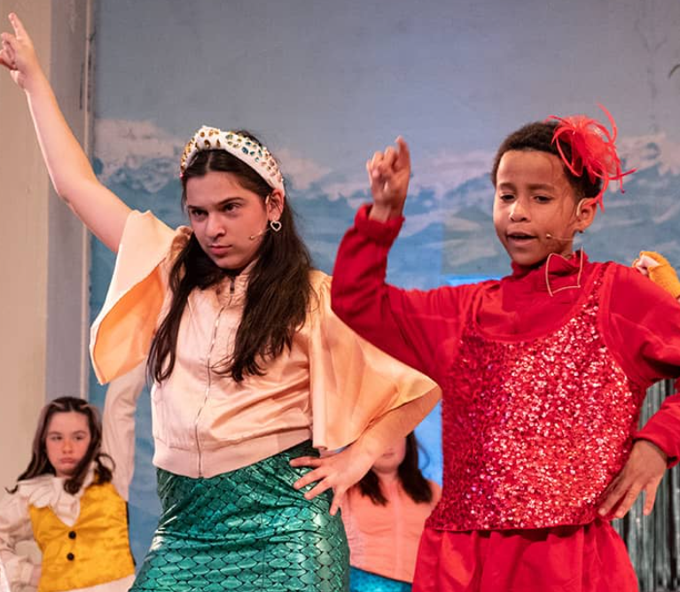 Two children on stage perform a dance, one in a mermaid costume with a headband and the other in a red sequined outfit with a feather. Members of the Youth Vocal Ensemble sit in the background, watching the lively performance unfold.