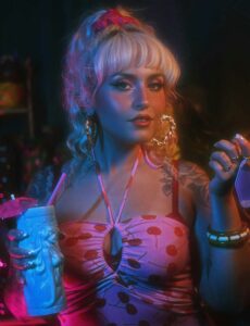 A woman with platinum blonde hair in a high ponytail wears a cherry-patterned dress and large hoop earrings, holding a drink in a ceramic tiki mug with a pink umbrella—an iconic scene from CCE Nightlife under colorful lighting.