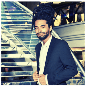 A man with dark dreadlocks and a short beard wears a dark suit and white shirt, standing in front of a modern staircase at Sevenings in Essex County. He has a calm expression and is buttoning his jacket.
