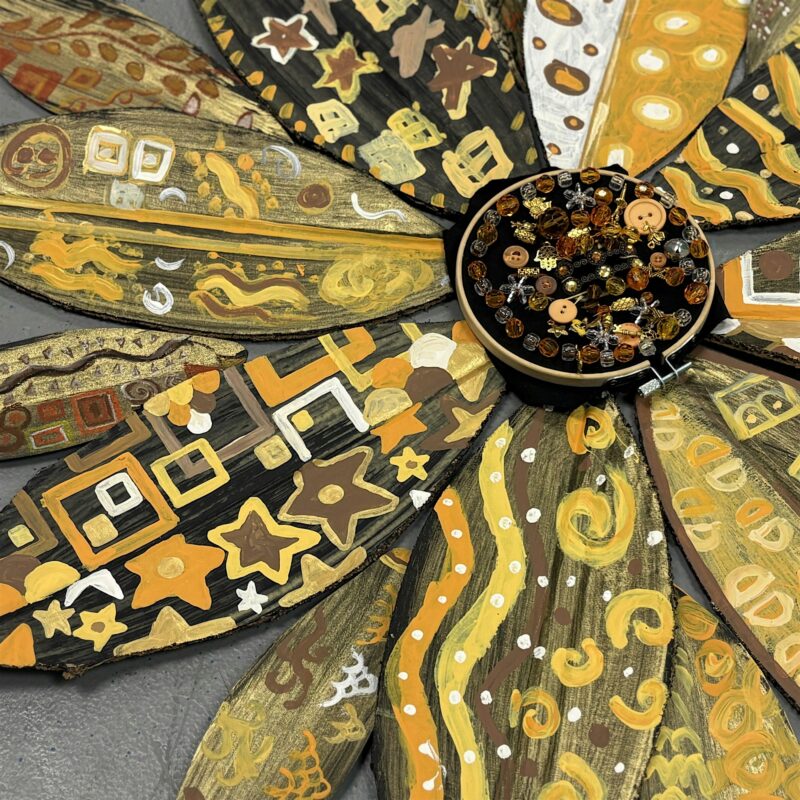 A close-up of a handcrafted decorative flower with painted petals featuring geometric, star, and swirl patterns in shades of brown, yellow, and orange. The center is filled with assorted beads and buttons.