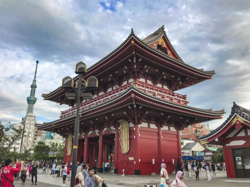 A large red traditional Japanese temple gate stands under a cloudy sky, with people walking around. The Tokyo Skytree tower is visible in the background to the left.