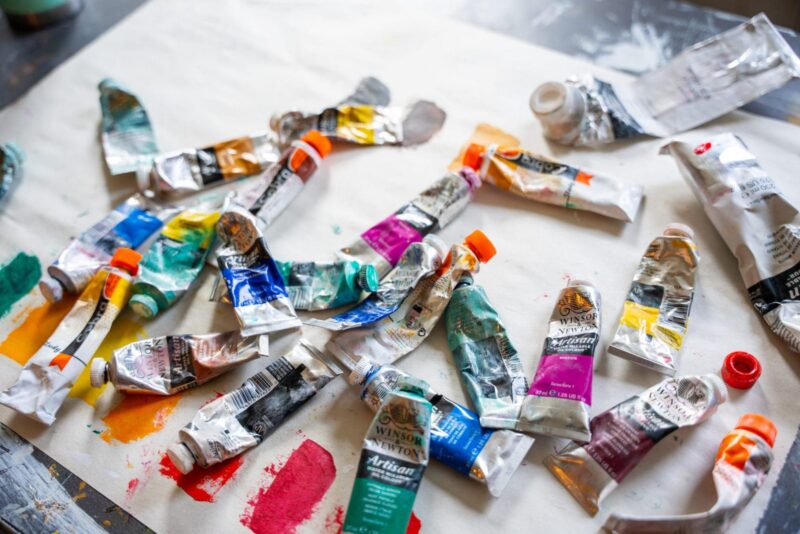 Several used and partially squeezed tubes of Winsor & Newton Artisan oil paint in various colors are scattered on a white canvas, with some paint marks and caps visible.