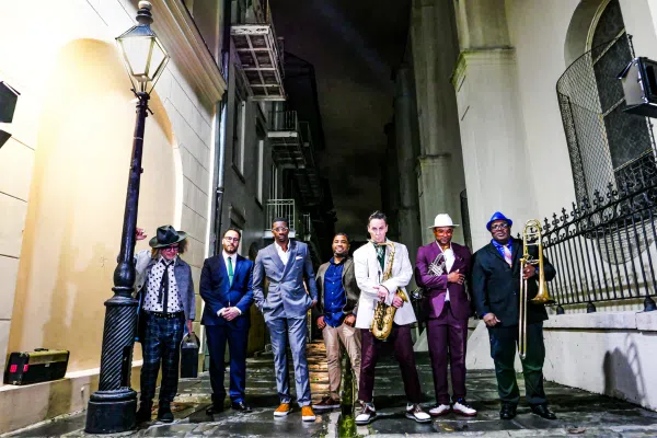 Seven men stand in an alley at night, dressed in suits and hats. Some hold musical instruments, including a saxophone and trombone, evoking the spirit of a New Orleans jazz band. A streetlamp and buildings complete the lively scene.