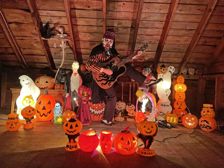 A person in a skeleton costume and face paint plays guitar in an attic decorated with numerous lit jack-o’-lanterns, Halloween figures, props, and a Dracula figure—perfect inspiration for your next spooky Drink n Draw session.