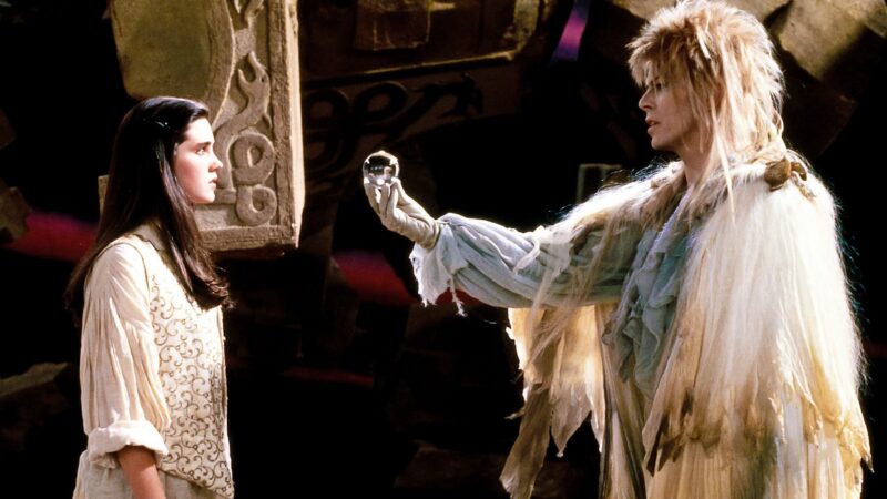 A young woman in a white outfit stands facing a figure with wild blond hair and a feathered cloak, who is holding a glass orb in an outstretched gloved hand. They stand in a dark, ornate setting.