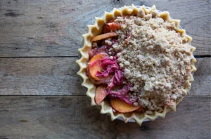 An unbaked fruit pie with a crimped crust sits on a wooden surface, ready for a Sevenings gathering in Essex County. The sliced fruit filling peeks through the crumbly streusel topping, promising a delightful treat.