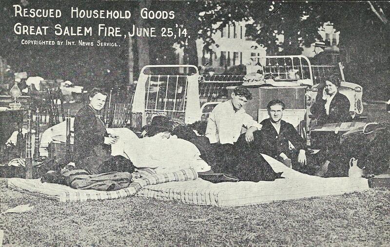 A black-and-white photo shows people sitting and lying on mattresses and bedding outdoors, surrounded by rescued household goods after the Salem Fire event on June 25, 1914. Pieces of furniture are visible in the background, marking Salem Fire history.