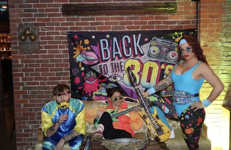Three people in colorful, eclectic costumes pose in front of a “Back to the 80’s” banner set against a brick wall. One person sits on a couch with a mustache prop, another lounges with green sunglasses, and the third stands holding a toy guitar.