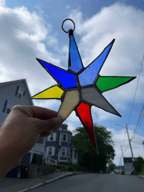 A hand holds up a stained glass star with blue, green, yellow, white, and red points against a partly cloudy sky and a residential street with houses and trees.