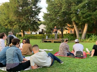 People sit on blankets on a grassy lawn watching a musician perform on a small stage outdoors, surrounded by trees and residential buildings in the background.