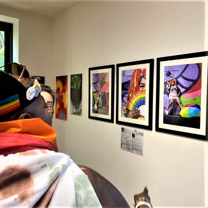 Two people view a wall display of colorful framed photographs and artwork, including images of people holding or surrounded by rainbow-themed items, in a brightly lit gallery space.