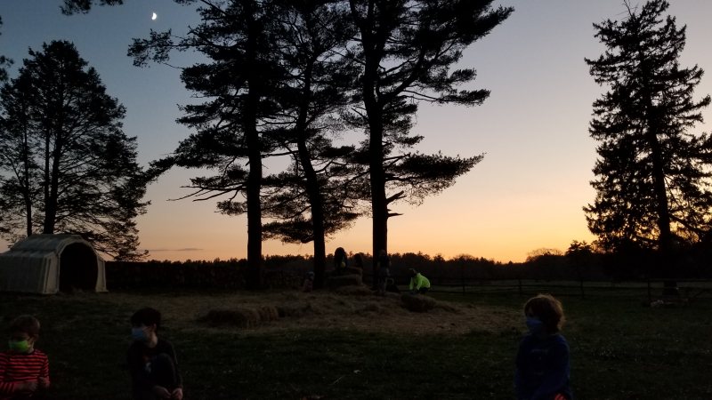 A group of children wearing masks sit and play near hay bales under tall trees at dusk, with a crescent moon and a small shelter visible in the background.