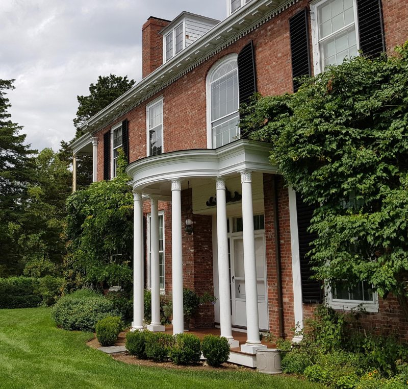 A large brick house with white columns, black shutters, arched windows, and ivy growing on the exterior, surrounded by green grass and shrubs.