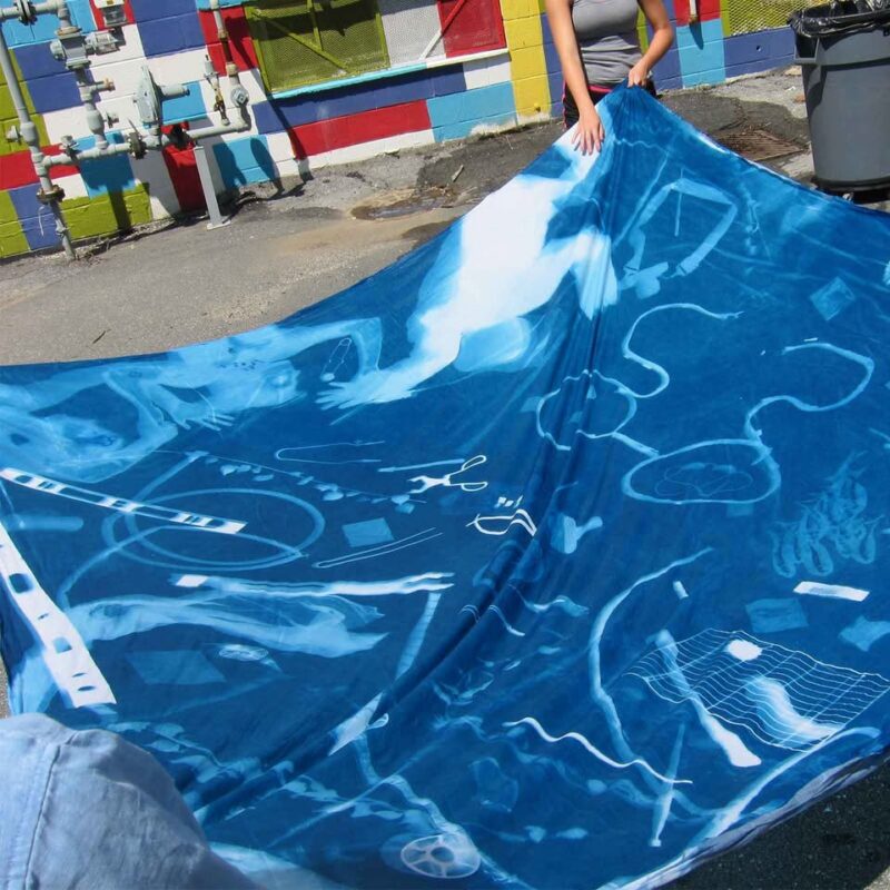 Two people hold a large blue fabric featuring white, abstract silhouettes reminiscent of cyanotypes outdoors. A colorful, tiled wall and utility pipes form the backdrop, reflecting creative photography and playful inspiration from Creative Foundations.