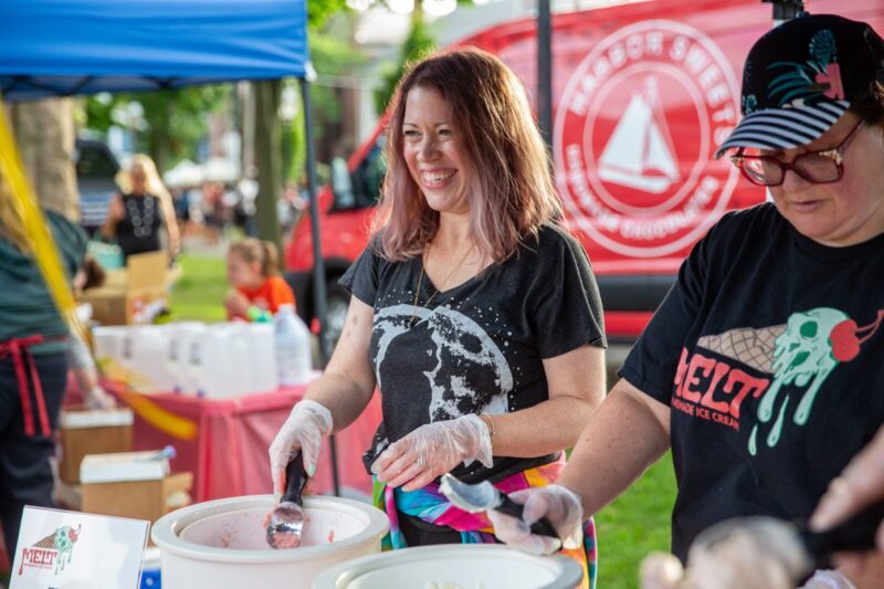 Two women wearing gloves serve ice cream from large containers at an outdoor event. One is smiling, and both are under a blue canopy. A red Melt ice cream truck and people are visible in the background.