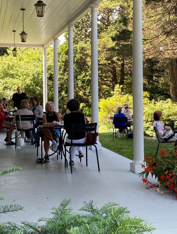 People are sitting at tables on a covered porch with white columns, talking and eating. More people are seated on the grass nearby. The scene is surrounded by trees and greenery on a sunny day.