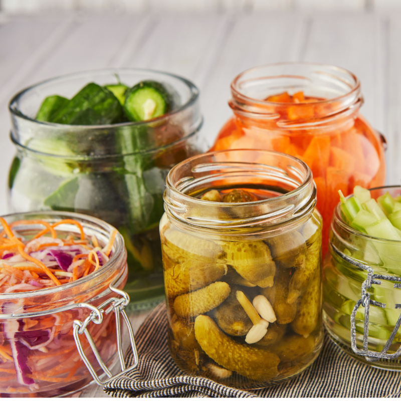 Five glass jars filled with assorted pickled and fresh vegetables, including cucumbers, carrots, cabbage with red onion, and gherkins, are arranged on a striped cloth with a light wooden background.