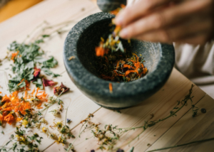 A hand places dried flowers and herbs into a stone mortar on a wooden surface, with additional dried botanicals scattered around—a serene Sevenings moment inspired by the natural beauty of Essex County.