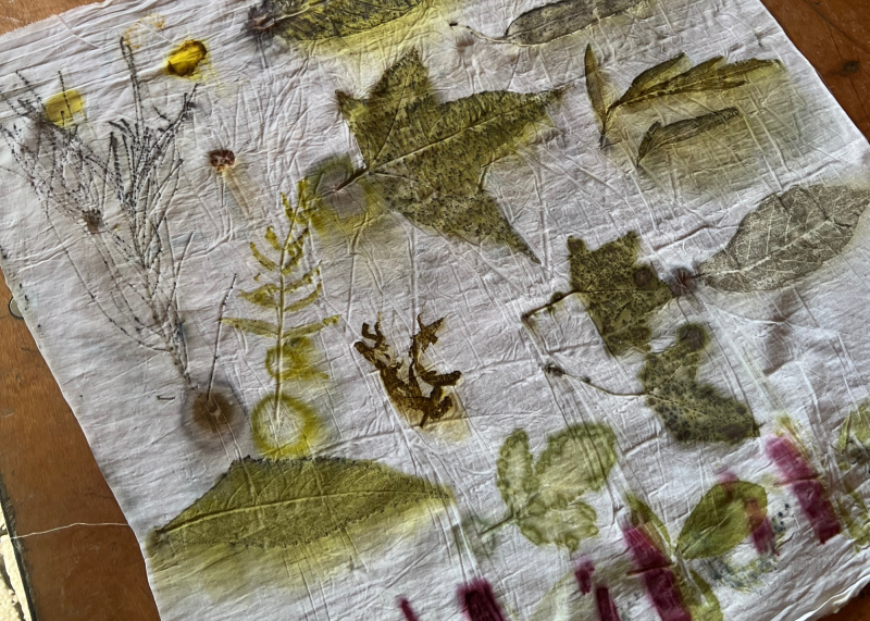 A piece of white fabric lies on a wooden surface, decorated with various natural leaf and plant imprints in green, yellow, brown, and purple shades. The designs appear organic and unevenly spaced.