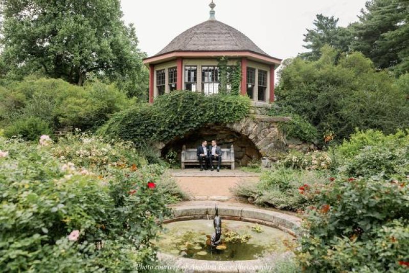 A round gazebo with a shingled roof stands above a stone alcove and bench, surrounded by lush greenery and flowering bushes. Two people sit on the bench. A small fountain with a statue is in the foreground.
