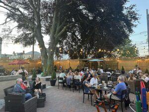 Outdoor restaurant patio in Essex County with people dining and chatting under string lights and a large tree. The area, perfect for Sevenings, is fenced with wooden panels, small plants, umbrellas, and an inviting early evening ambience.