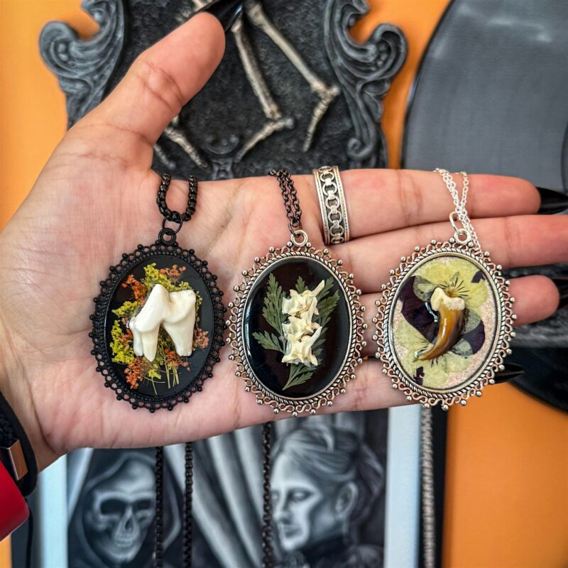 A hand holds three ornate necklaces, each with a pendant containing preserved teeth or fangs, set against a black background with botanical elements. Gothic-style artwork and decor are visible in the background.