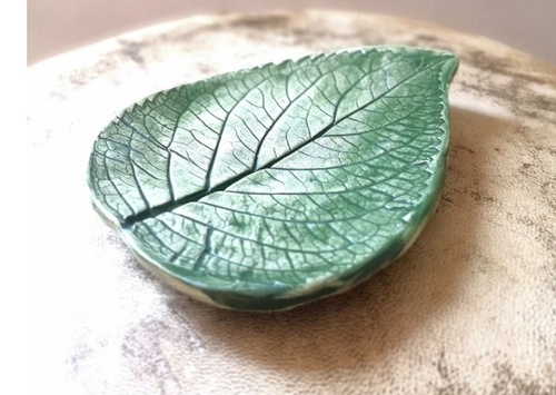 A small, green ceramic dish shaped and textured like a leaf rests on a light, textured surface. The dish features detailed vein patterns and a glossy finish.