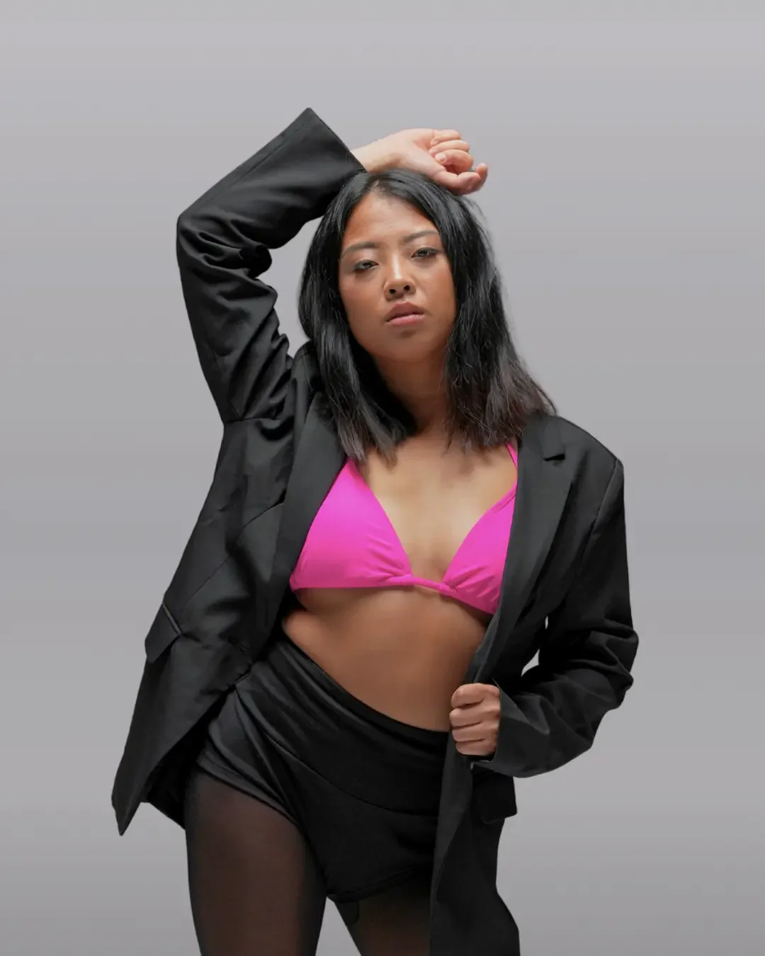 A woman stands against a plain gray background wearing a black blazer, a bright pink bikini top, black shorts, and sheer tights. One arm is raised with her hand resting on her head. She looks directly at the camera.
