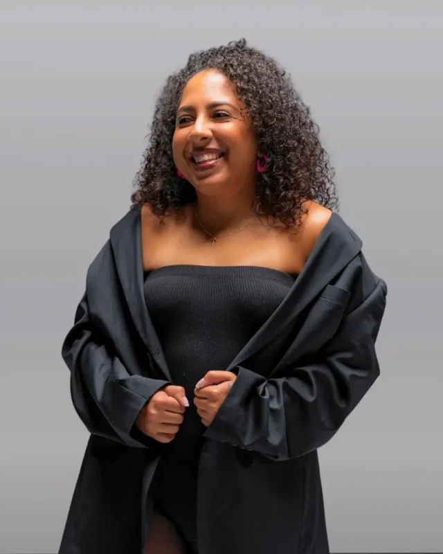 A woman with curly hair smiles while standing against a plain gray background. She is wearing a black off-the-shoulder top or dress and a loose black jacket, holding the jacket closed with both hands.