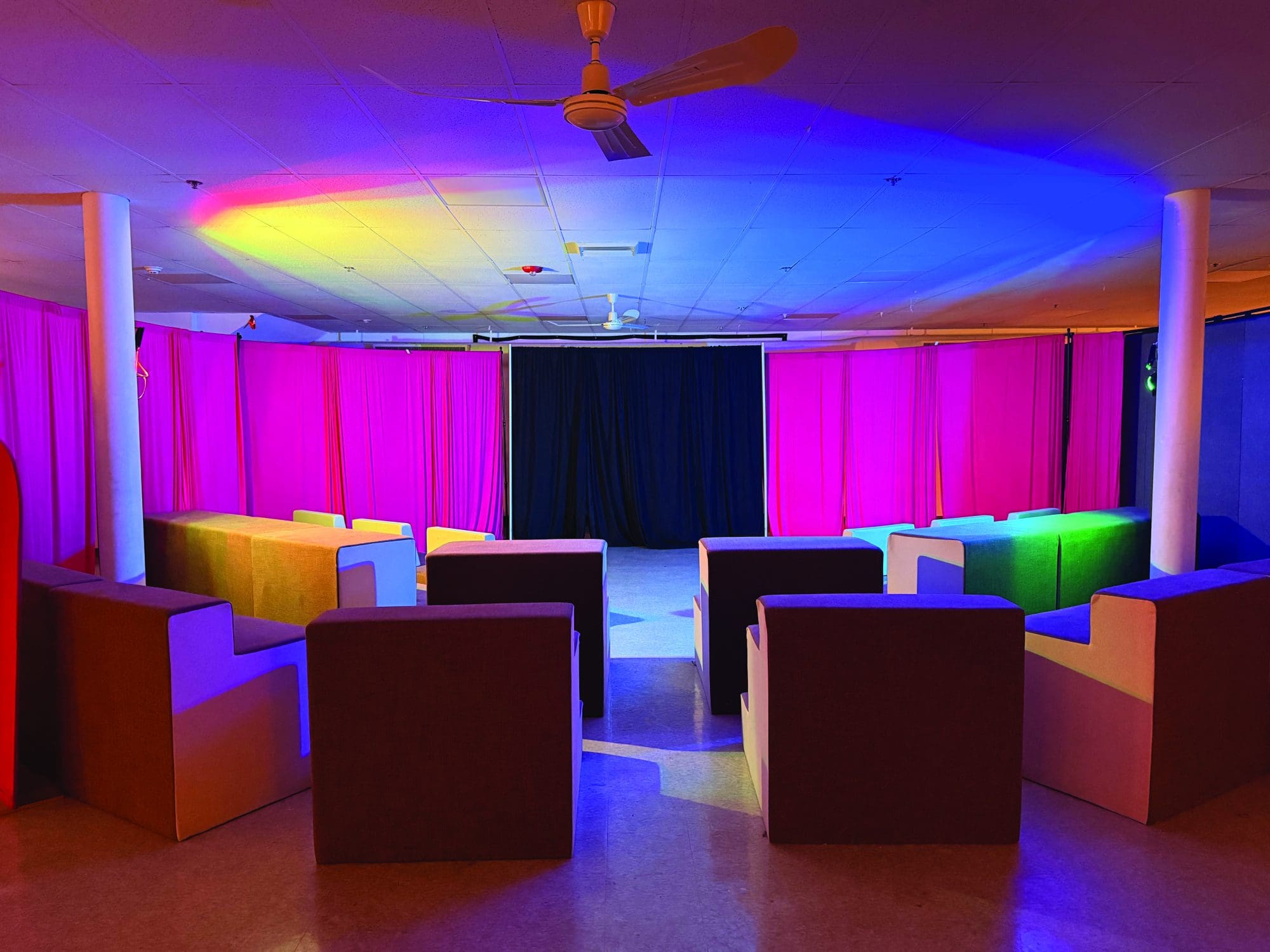 A room with armchairs arranged in a semicircle faces a stage with black curtains. Pink and blue lights illuminate the ceiling and walls, set for Bower Shows at the 2026 Beverly Fringe Festival, with a ceiling fan visible above.