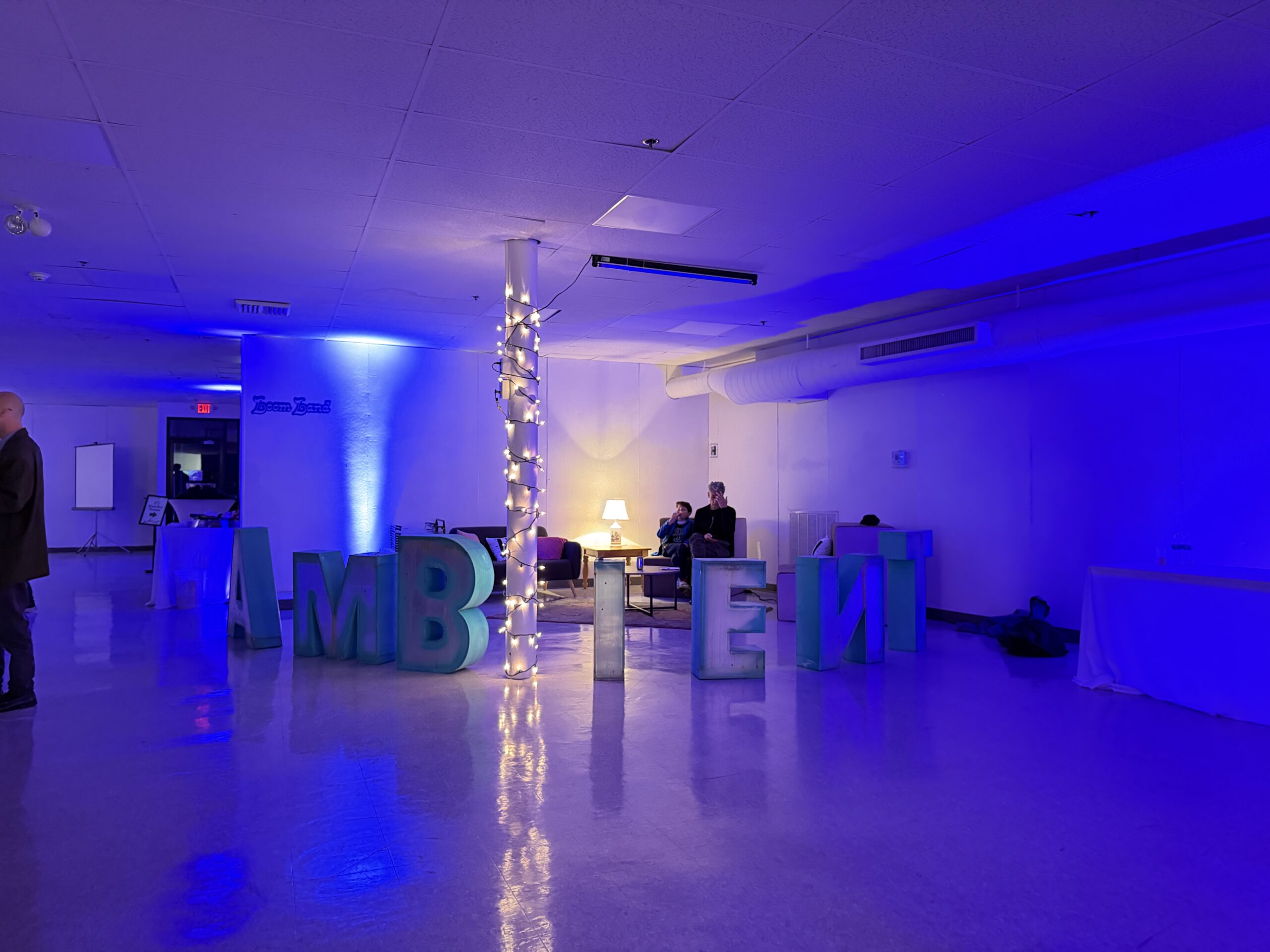 A large room with blue lighting at The Bower Shows, oversized letters spelling "AMBITION," two people on a couch near a lit lamp, and a pole wrapped in string lights—a scene from the 2026 Beverly Fringe Festival.