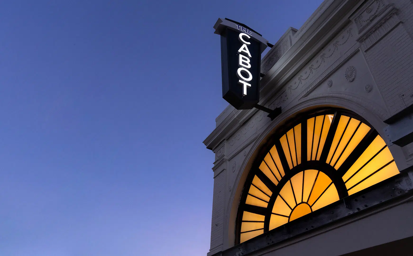 A building facade with an arched window illuminated in yellow light, beneath a vertical sign reading "THE CABOT" against a clear evening sky—perfect for Member Tours seeking architectural charm.