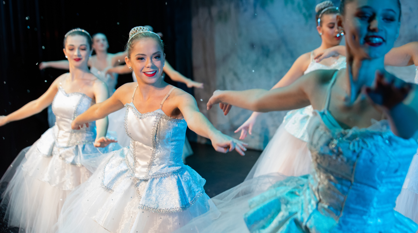 Four ballet dancers in white costumes and tiaras perform on stage with their arms extended, smiling and looking in different directions. The softly lit blue and white background highlights this enchanting Performing Arts Academy showcase.