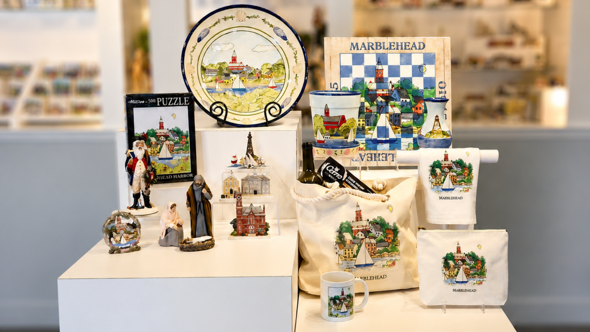 A display of Marblehead-themed souvenirs by Hestia Creations, including a puzzle, decorative plate, figurines, mug, tote bags, dish towels, and postcards—all showcasing handcrafted artistry and local landmarks.