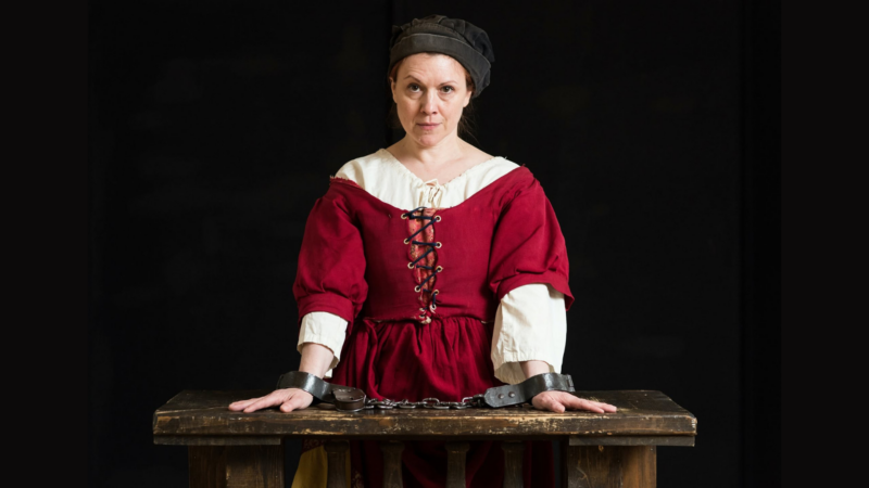 A woman in historical clothing stands behind a wooden table with her hands in metal shackles. She wears a red dress with a white blouse underneath and a black cap, bringing History Alive against a dark background.