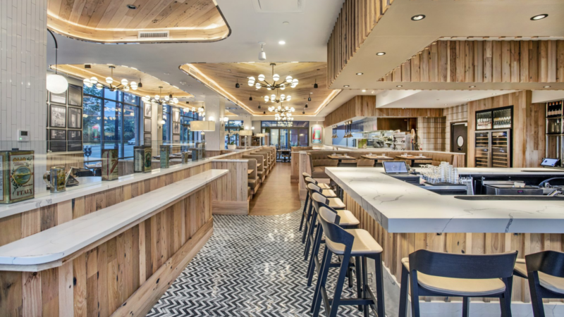 A modern restaurant interior by JJ Welch Construction features wooden paneling, marble countertops, bar seating, chandeliers, patterned tile and wood floors, with large windows filling the space with natural light. Tables and booths are thoughtfully arranged throughout.