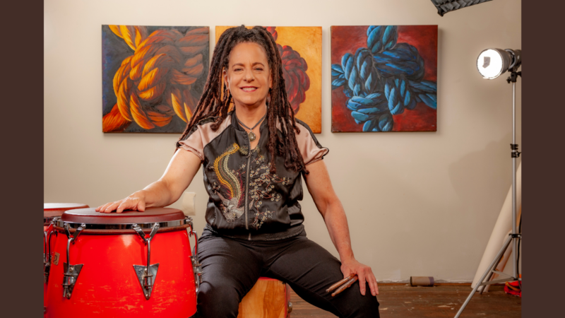 A woman sits on a wooden box with one hand resting on red conga drums. She holds drumsticks and smiles at the camera. Behind her are three colorful abstract paintings on the wall and a photography light to the right.