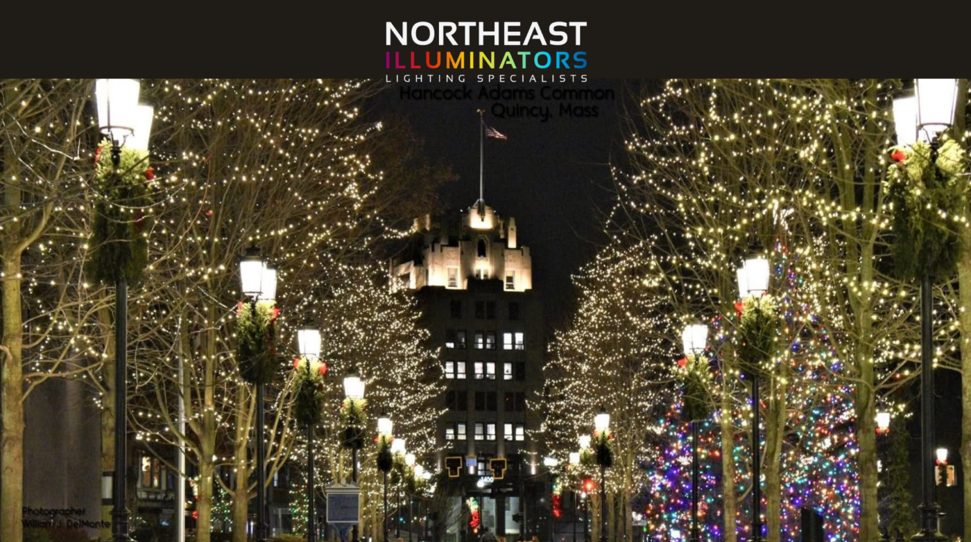 A nighttime scene of Hancock Adams Common in Quincy, Massachusetts, featuring trees and lampposts decorated by Northeast Illuminators with string lights and wreaths, and a lit building and holiday tree glowing in the background.